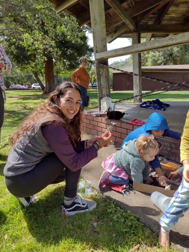 Esme, a young latinx woman, holding a feather and kneeling next to playing toddlers